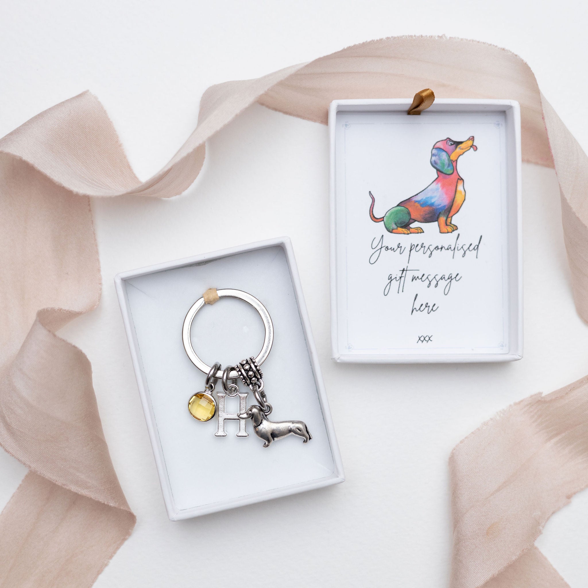 Personalised dachshund keyring with silver colour sausage dog charm, birthstone, and initial. Photographed in a gift box, with the lid showing an illustration of a dachshund and a gift note