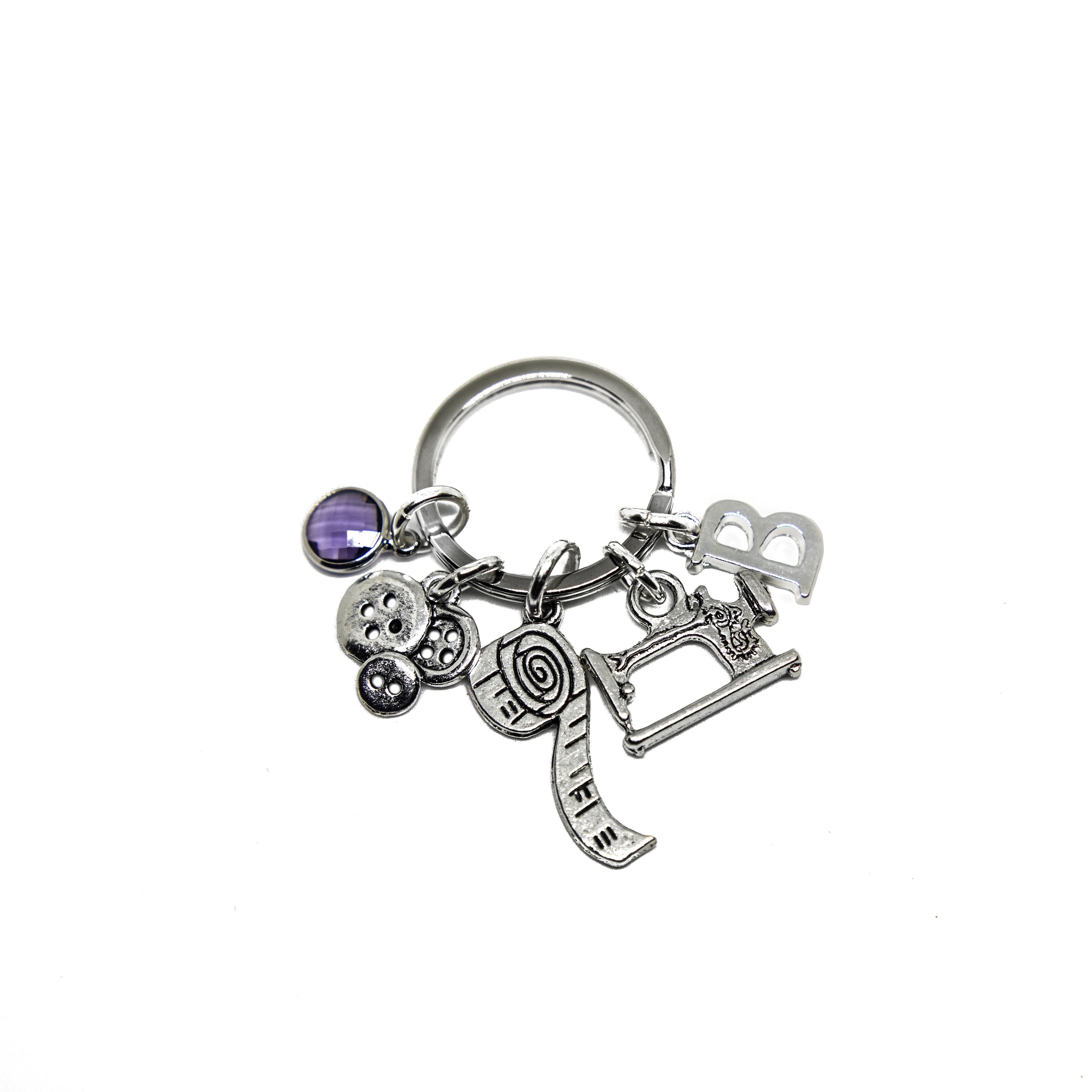 A silver keyring with multiple charms, including a sewing machine, buttons, and a personalized letter charm, with a small purple birthstone.