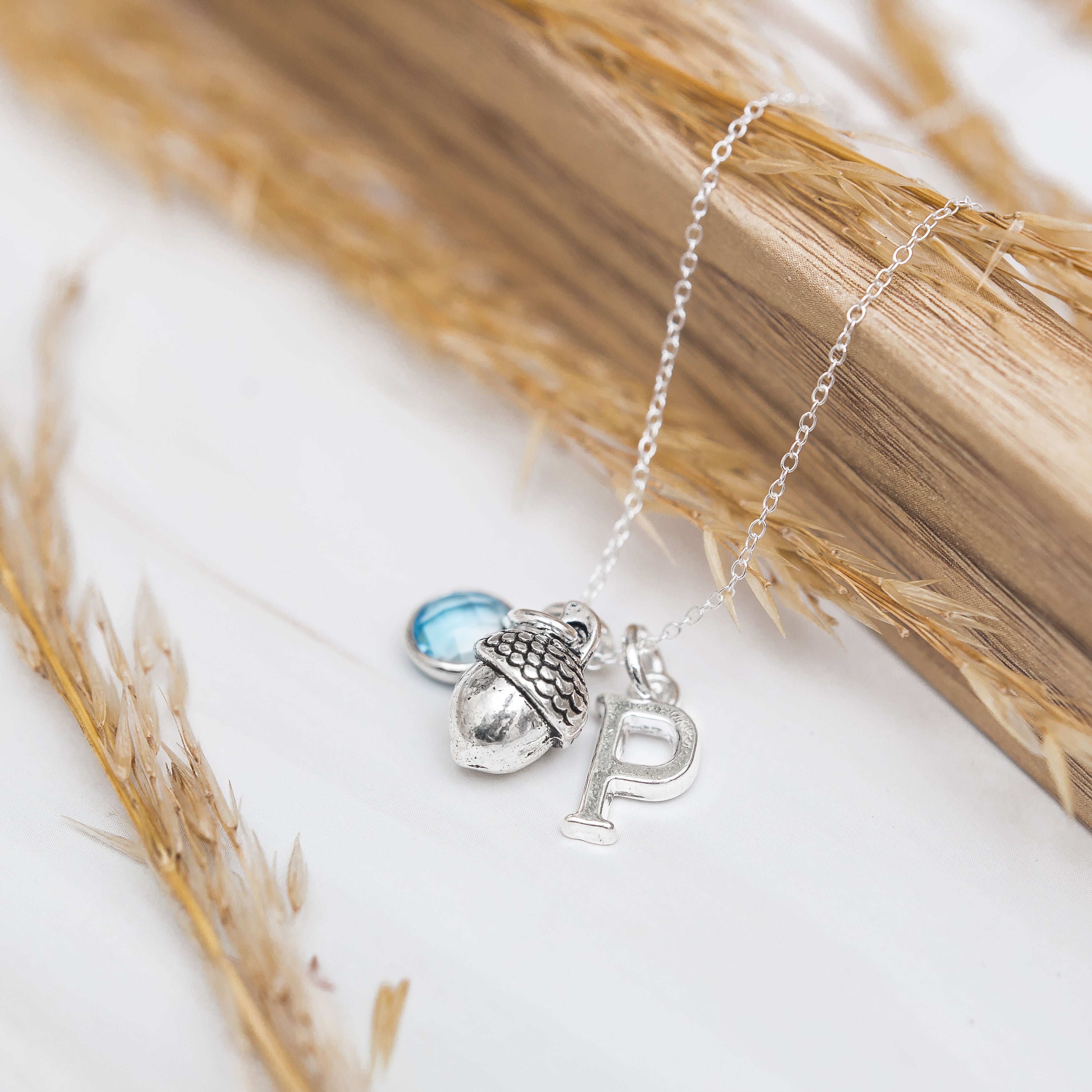 Customised pendant necklace with a trio of charms on a dainty trace chain. The necklace has a birthstone charm, then an acorn charm and then a letter charm.