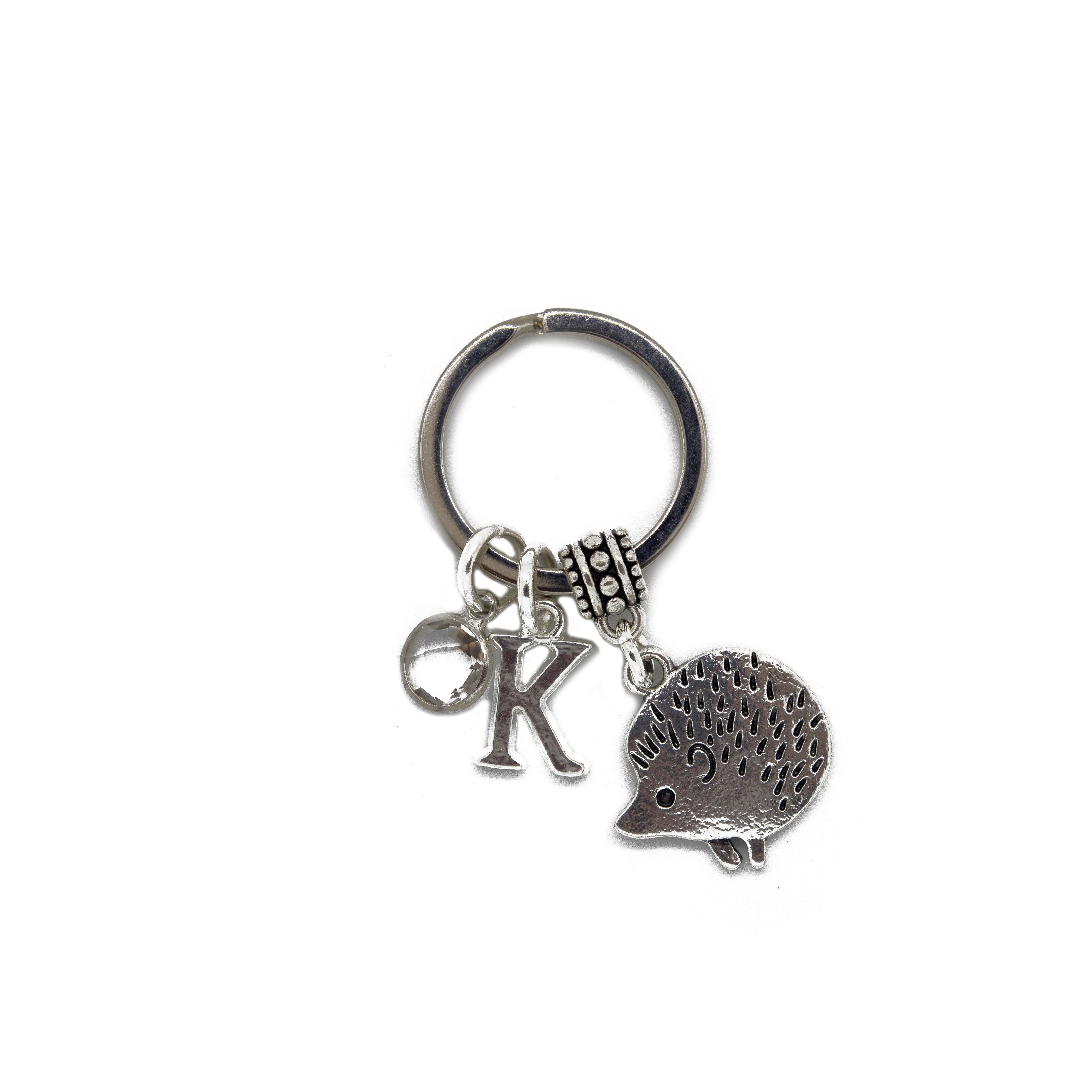 A silver-colored metal alloy keyring featuring a charm shaped like a hedgehog, with a letter charm and a birthstone charm also attached to the keyring.